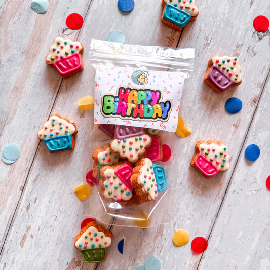 Mini cupcake-shaped dog treats with colorful toppings, packaged in a clear bag with a 'Happy Birthday' label, displayed on a wooden surface with confetti scattered around.